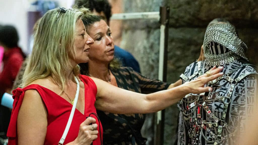 Due visitatrici indicano i dettagli di un abito-scultura in metallo su un manichino durante una mostra.